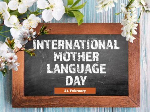 A wooden-framed chalkboard with the text 'INTERNATIONAL MOTHER LANGUAGE DAY' written in white chalk. Below the main text, a small orange rectangle contains the date '21 February.' The chalkboard is surrounded by white and light pink flowers on a light blue wooden background.