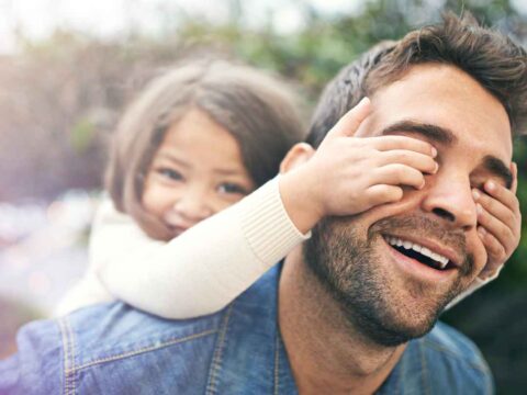 Father joyfully carrying his daughter on his back as she playfully covers his eyes with her hands. This features a perfect image for celebrating Daughters Day.